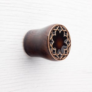 May include: A dark brown wooden ear plug with a decorative geometric design on the face. The design features a series of interconnected shapes filled with a dark, glossy material. The plug has a smooth, rounded shape.