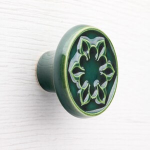 May include: A round ceramic cabinet knob featuring a glossy dark green finish with a lighter green and white floral-inspired pattern centered on the face. The knob has a smooth, reflective surface and a cylindrical base extending from the back.