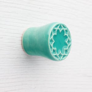 Puede incluir: Pomo de armario de cerámica turquesa con un diseño decorativo de estrella. El pomo tiene forma cilíndrica con un extremo ligeramente ensanchado y un acabado liso y brillante. El diseño es un patrón geométrico.