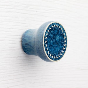 Puede incluir: Un pomo de armario de cerámica azul con un centro azul oscuro texturizado y un borde blanco punteado. El pomo tiene forma redondeada y un efecto veteado azul claro en el vástago.