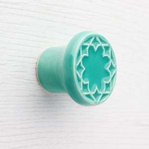 Pode incluir: Puxador de armário de cerâmica turquesa com um design de estrela. O puxador é redondo com uma base cilíndrica e é montado em uma superfície de madeira branca. O padrão de estrela é embutido na face do puxador.