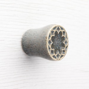 Könnte beinhalten: Grauer Keramik-Schrankknopf mit floralem Design auf der Vorderseite. Der Knopf hat eine zylindrische Form mit einem dekorativen Muster in dunklerer Blaufärbung. Der Knopf ist etwa 3,8 cm lang.