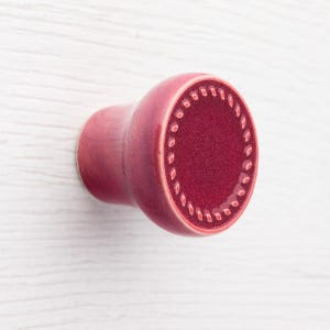 Op de afbeelding: Een close-up van een rode kastknop. De knop heeft een afgeronde vorm met een gestructureerd, cirkelvormig ontwerp op de voorkant. De knop is gemaakt van keramiek of een vergelijkbaar materiaal en is gemonteerd op een wit oppervlak.