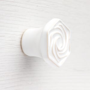 Op de afbeelding: Een witte keramische meubelknop met een zeshoekig voorvlak voorzien van een verhoogd, wervelend spiraalpatroon. De knop heeft een gladde, matte afwerking en een cilindrische basis die licht taps toeloopt naar het midden toe.