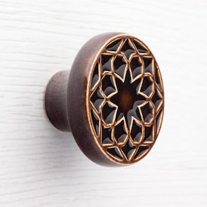 Könnte beinhalten: Brauner Keramik-Schrankknauf mit Sternmuster. Der Knauf hat eine dunkelbraune Oberfläche mit einem detaillierten geometrischen Muster. Der Knauf hat einen Durchmesser von ca. 5 cm und ist auf einem Holzsockel montiert.