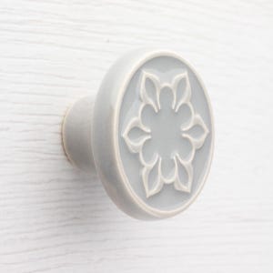 Puede incluir: Pomo de armario de cerámica gris claro con diseño floral. El pomo tiene forma redondeada con un diseño floral blanco en relieve en el centro. El fondo es de color gris claro.