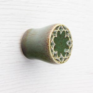 May include: A green and brown ceramic drawer pull with a decorative starburst design on the front. The pull is cylindrical with a flared end and is set against a white background.