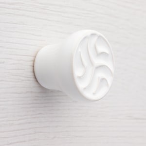 Może przedstawiać: Białe, okrągłe pokrętło do szafki z dekoracyjnym, wirującym wzorem na froncie. Pokrętło wykonane jest z litego materiału i zamontowane na jasnej drewnianej powierzchni. Prosty, ale elegancki element okuć.