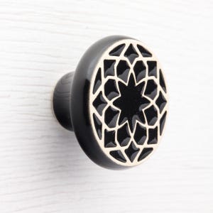 Może przedstawiać: Czarny ceramiczny uchwyt do szafki z geometrycznym wzorem gwiazdy. Uchwyt ma błyszczące wykończenie i kontrastującą kremową obwódkę wokół wzoru gwiazdy. Okucia są zamontowane na białej powierzchni.