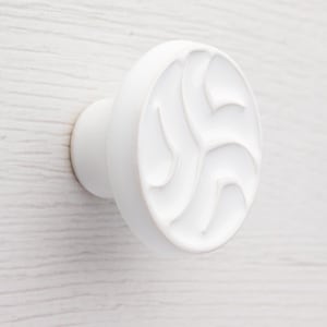 Puede incluir: Perilla de armario redonda blanca con un diseño decorativo de remolino en relieve. La perilla tiene un acabado liso y brillante. La perilla está montada sobre una superficie blanca. Hecho de material sólido.