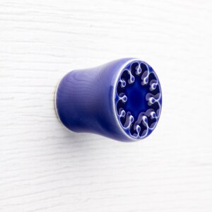 Può includere: Pomello per armadio cilindrico in ceramica blu con un design decorativo sulla parte anteriore. Il pomello ha un corpo liscio e arrotondato e una base bianca a contrasto. Il design presenta una serie di forme stilizzate interconnesse.