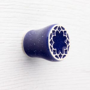 May include: A dark blue ceramic cabinet knob with a white starburst design on the face. The knob has a speckled finish and is mounted on a white surface. This decorative hardware adds a unique touch to furniture.