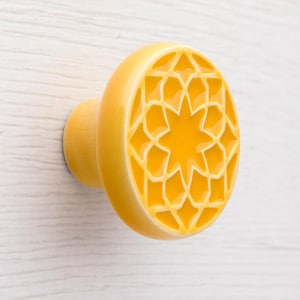 May include: A yellow cabinet knob with a geometric starburst design. The knob is round with a flat back and a decorative front. The design is a series of interconnected shapes, creating a star in the center.