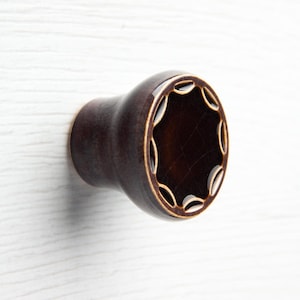 Può includere: Pomello per armadio in ceramica marrone scuro con bordo smerlato decorativo. Il pomello ha una finitura lucida ed è montato su una superficie di legno bianco. Il design presenta una serie di intagli ovali sul bordo.