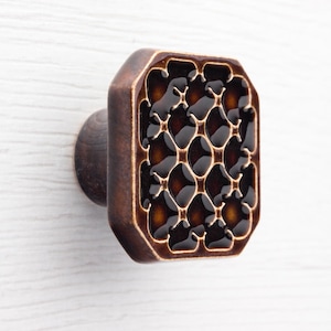 Può includere: Pomello decorativo per mobili con una faccia rettangolare marrone scuro con un motivo di cerchi interconnessi. Il pomello ha un bordo color bronzo e una base rotonda marrone scuro per il montaggio. Il design ricorda un favo.