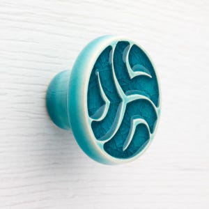 Puede incluir: Pomo de armario de cerámica turquesa con diseño circular. El pomo presenta un patrón abstracto en relieve en un tono más oscuro de turquesa, con una textura craquelada. Los bordes son de color turquesa más claro.
