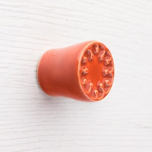 Op de afbeelding: Een cilindrische, oranjerode oorplug met een decoratief, wervelend patroon aan het uitlopende uiteinde. De plug heeft een gladde, gepolijste afwerking en een licht getextureerd uiterlijk op het lichaam. Het ontwerp is symmetrisch.