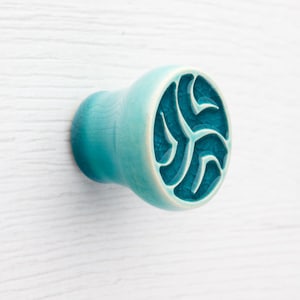May include: A turquoise ceramic cabinet knob with a decorative carved design. The knob has a rounded shape with a circular face featuring an intricate pattern. The knob is mounted on a white wooden surface.