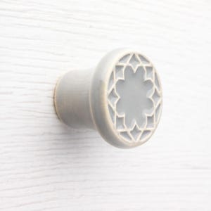 May include: A light grey cabinet knob with a cylindrical base and a circular face featuring a recessed star-shaped pattern. The surface of the knob has a smooth, glossy finish, and the geometric design consists of a central star surrounded by diamond-shaped cutouts.