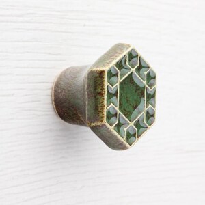 May include: A ceramic cabinet knob with a hexagonal face. The knob has a dark green and grey geometric design on the face and a brown stem. The knob is designed to be mounted on a cabinet or drawer.
