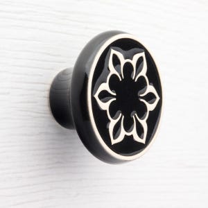 Op de afbeelding: Zwarte en witte keramische kastknop met een bloemenontwerp. De knop heeft een glanzende afwerking en een ovale vorm. Het bloemenontwerp is wit tegen een zwarte achtergrond.