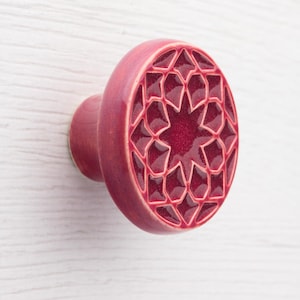 Może przedstawiać: Okrągła, dekoracyjna gałka do szafki w głębokim odcieniu czerwieni. Gałka ma geometryczny wzór gwiazdy wycięty na jej powierzchni. Gałka jest wykonana z ceramiki i ma gładkie, błyszczące wykończenie. Trzpień ma pasujący czerwony kolor.