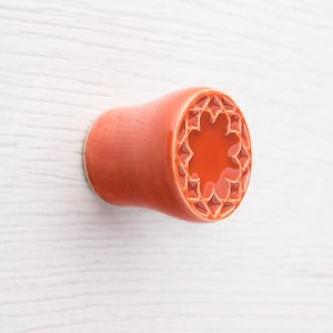 May include: A cylindrical, orange-colored ear plug with a decorative, star-shaped design on the end. The plug is made of ceramic and has a smooth, glossy finish. The design is carved into the end of the plug.