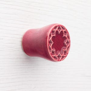 May include: A maroon-colored ear plug with a decorative, star-shaped design on the end. The plug is cylindrical with a flared end and is set against a white wooden background.