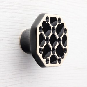 Op de afbeelding: Zwarte en crèmekleurige achthoekige kastknop met een patroon van zwarte vormen. De knop heeft een afgeronde basis en is gemonteerd op een wit houten oppervlak.