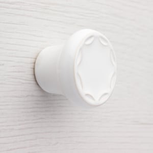 Peut inclure: Bouton de meuble en céramique blanche avec une face circulaire et une base cylindrique. La face présente un motif de feuilles répétées sur le bord. Le bouton est posé sur un fond clair en imitation bois.