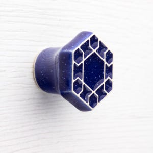 Puede incluir: Pomo de armario de cerámica azul con diseño geométrico. El pomo tiene forma hexagonal con contorno blanco y acabado moteado. El diseño presenta una serie de formas interconectadas.
