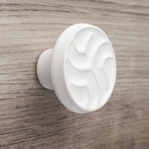 Könnte beinhalten: Ein weißer, runder Schrankknauf mit einem dekorativen Wirbeldesign. Der Knauf ist auf einer Holzoberfläche mit einem hellbraunen Maserungsmuster montiert. Diese Hardware ist für Schubladen und Schränke geeignet.