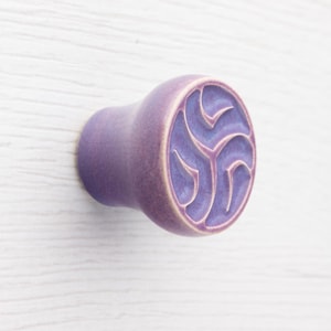 Może przedstawiać: Lawendowy ceramiczny uchwyt do szafki z okrągłym wzorem. Uchwyt ma zaokrąglony kształt z wyrzeźbionym, trójdzielnym wzorem spiralnym z przodu. Kolor to miękki fiolet z lekko teksturowanym wykończeniem.