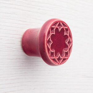 Puede incluir: Un tapón para el oído de cerámica de color burdeos con un diseño de estrella. El tapón tiene una cara redondeada con un patrón geométrico y un vástago cilíndrico. El fondo es una superficie blanca con vetas de madera.