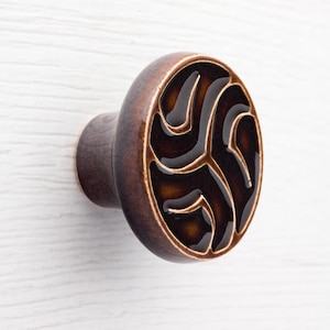 Może przedstawiać: Brązowy ceramiczny uchwyt do szafki o okrągłym wzorze. Uchwyt ma ciemnobrązowy, błyszczący środek z wirującym wzorem, otoczony jaśniejszą brązową obwódką. Zamontowany na białej drewnianej powierzchni.