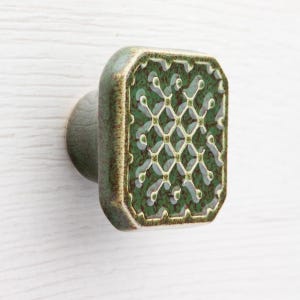Puede incluir: Pomo de armario cuadrado de cerámica verde con un patrón de celosía decorativo. El pomo tiene un aspecto envejecido y vintage y está montado sobre una superficie de color claro. El pomo es un elemento de ferretería decorativo.