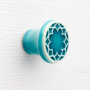 Puede incluir: Pomo de cerámica turquesa con borde blanco y diseño floral geométrico. El pomo tiene una textura craquelada y está montado sobre una superficie blanca. Este herraje decorativo es adecuado para cajones y armarios.