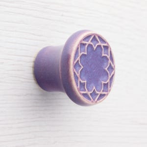 Puede incluir: Un tirador de armario de cerámica morada con un diseño geométrico de estrella. El tirador tiene forma redondeada con una parte trasera plana para el montaje. El patrón de estrella está grabado en la superficie.