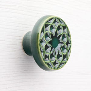 Puede incluir: Un pomo de armario de cerámica verde con un diseño de estrella. El pomo tiene forma redondeada con un centro verde oscuro y un borde verde más claro. El patrón de estrella es una mezcla de verde y blanco.