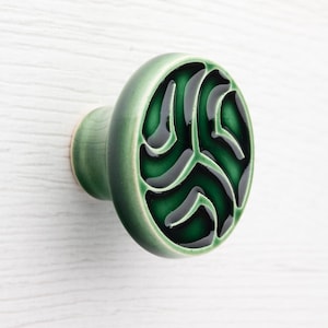 Puede incluir: Un pomo de cerámica verde con un diseño circular. El pomo presenta un acabado brillante con un patrón verde oscuro sobre una base verde más clara. El pomo está montado sobre un vástago verde claro.