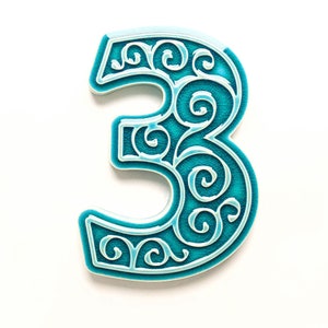 May include: A turquoise blue ceramic number three with a white swirl design. The number is a decorative accent for a home.
