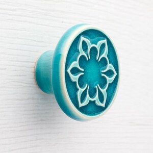 Può includere: Pomello per armadio in ceramica turchese con un design floreale. Il pomello ha una faccia rotonda con un motivo floreale in rilievo in una tonalità più scura di turchese. I bordi sono turchese chiaro con un bordo bianco.