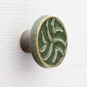 Op de afbeelding: Een keramische kastknop met een groen en bruin afwerking. De knop heeft een cirkelvormig ontwerp met een groen midden en een decoratief patroon. De knop is gemonteerd op een wit oppervlak.