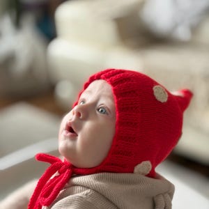 May include: A red knitted baby bonnet with white polka dots and long ties. The bonnet is worn by a baby wearing a beige hooded sweater. The baby has blue eyes and is looking upwards.