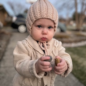 May include: A young child wearing a light pink knitted hat with a pointed top and a cream colored knitted cardigan. The child is holding two small rocks in their hands.
