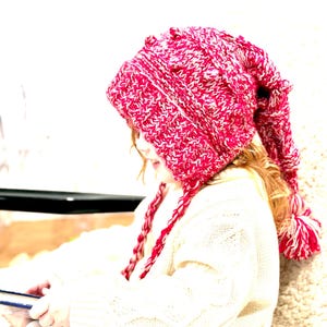 May include: A red and white knit hat with a long pointed end and a matching pom-pom. The hat has braided ties and is worn by a person wearing a cream-colored sweater.