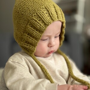 May include: A knitted olive green baby bonnet with ear flaps and ties. The bonnet has a pointed top and a textured knit pattern. The baby is wearing a light beige long-sleeved shirt.