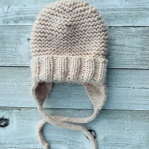 May include: A light beige knitted hat with ear flaps and ties. The hat is made of a soft, textured yarn and features a ribbed brim. The ear flaps extend down to the neck and have long ties for securing the hat. A cozy winter accessory.