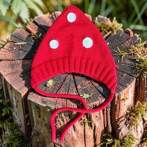 May include: A bright red knitted hat with a pointed top and a ribbed brim. The hat features three circular white felt spots on the front and includes two long red knitted ties attached to the sides for securing the hat under the chin.