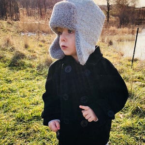 May include: A young child wearing a black coat and a gray fuzzy winter hat with ear flaps. The child is standing in a grassy field.